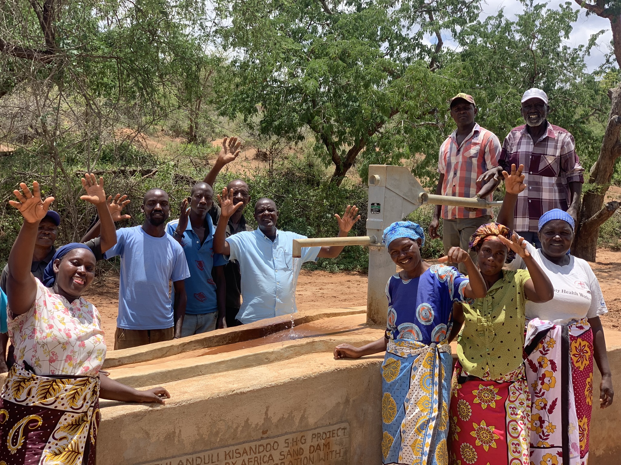 ten community members stand around raised water pump waving