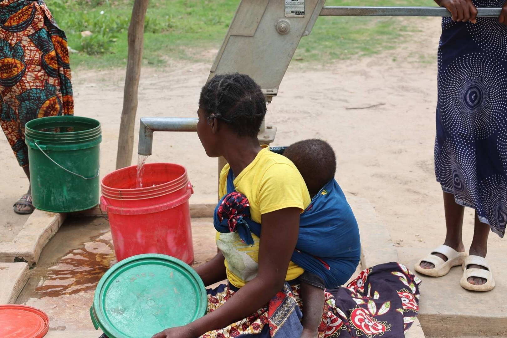 Woman with baby on back fills up water bucket from pump