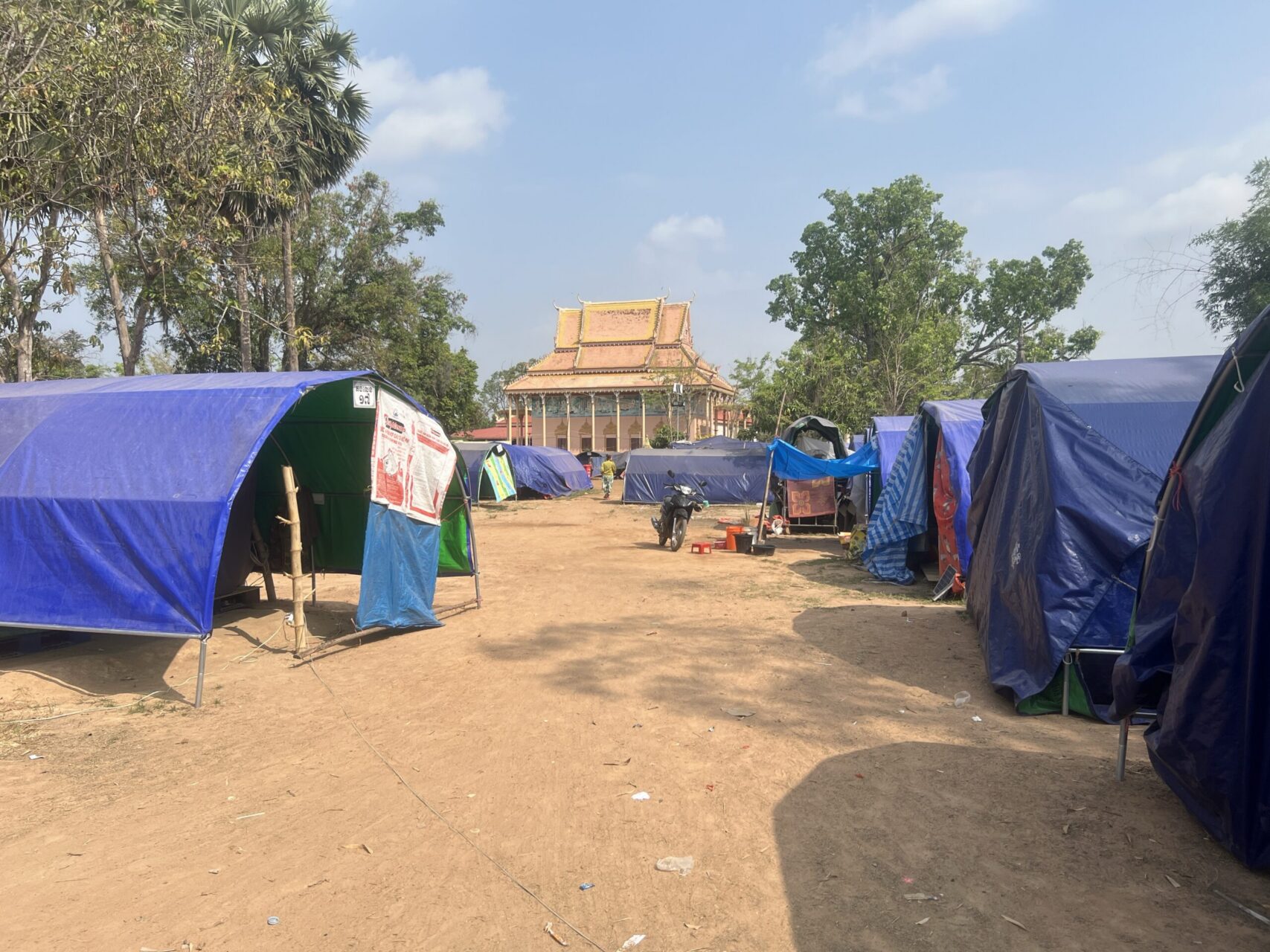 Blue tents are lined up either side of a sandy pathway, with a temple in the background