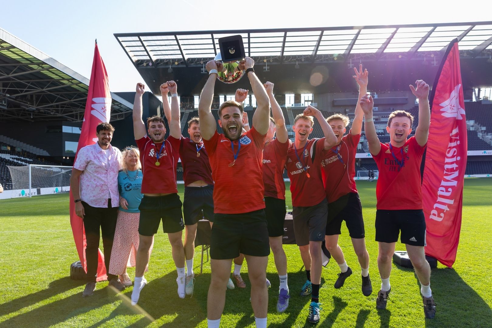 A group of men in red football tops jump into the air. The man at the front is holding a trophy
