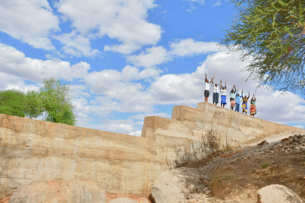 Seven people stand on top of a sand dam with their arms in the air